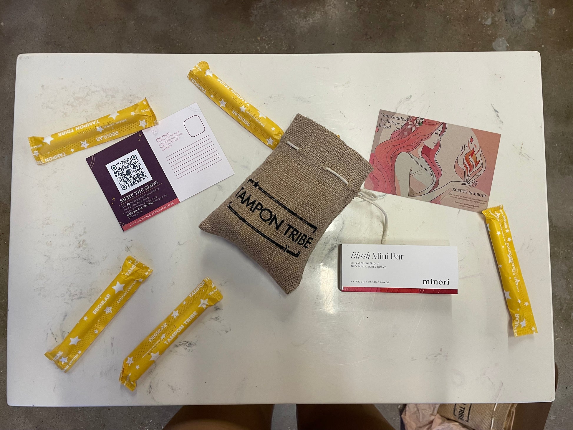 Assorted products including yellow bars, a brown bag labeled 'Tampon Tribe', and a white box on a white surface.